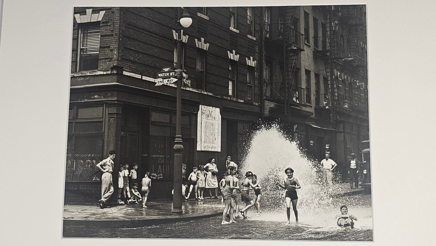 A black and white vintage street photograph print mounted with a wide white mat board, depicting children playing in a city street with buildings and a fountain spray.