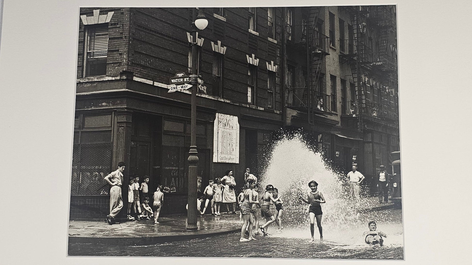 A black and white vintage street photograph print mounted with a wide white mat board, depicting children playing in a city street with buildings and a fountain spray.