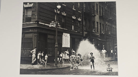 A black and white vintage street photograph print mounted with a wide white mat board, depicting children playing in a city street with buildings and a fountain spray.